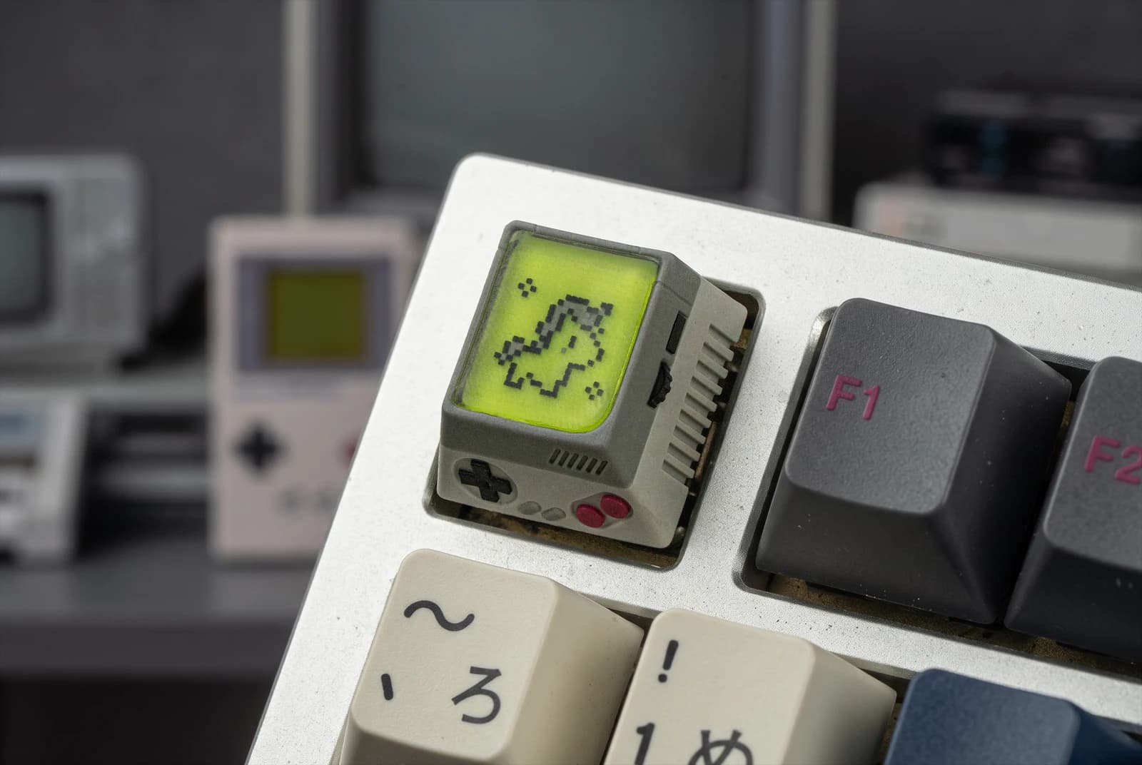 Close-up of the 1U R1 DMG Handheld artisan at Escape, showing a gray console-shaped body, dark directional pad, red buttons and yellow-green pixel-style screen.