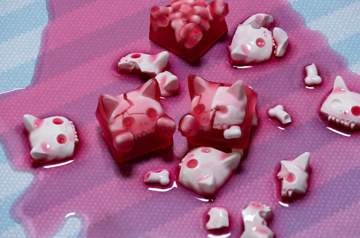 Translucent red Gelatinous Rouge cat artisan keycaps with pale skull details, surrounded by miniature skulls and bones on a pink and blue surface.