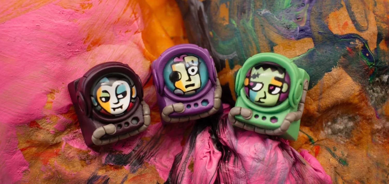 Vlad, Suzie and Frank Monster Mash Cadet artisan keycaps in burgundy, purple and green helmets, on a colourful painted background.
