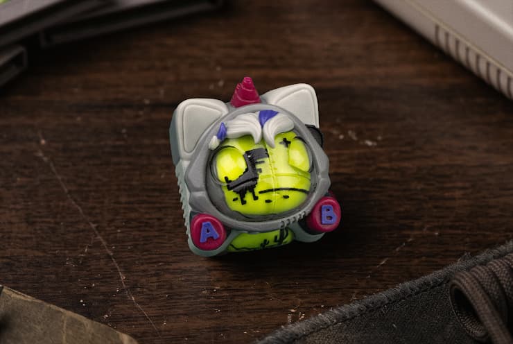 DMG Napicorn artisan keycap with a gray cat-eared shell, magenta horn and A/B buttons around a bright yellow-green feline face.